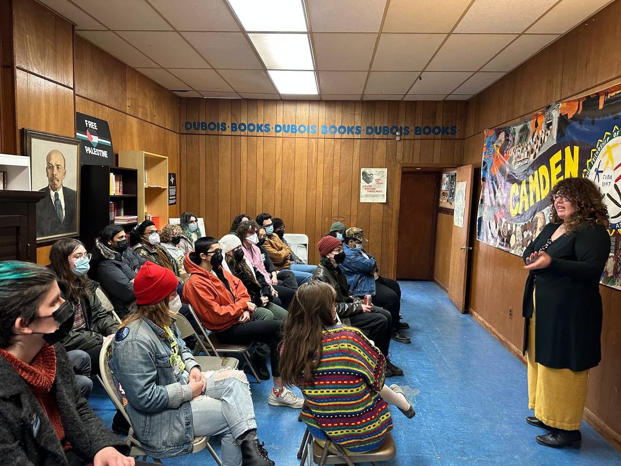 A photo of many people sitting around a room with a speaker addressing the crowd