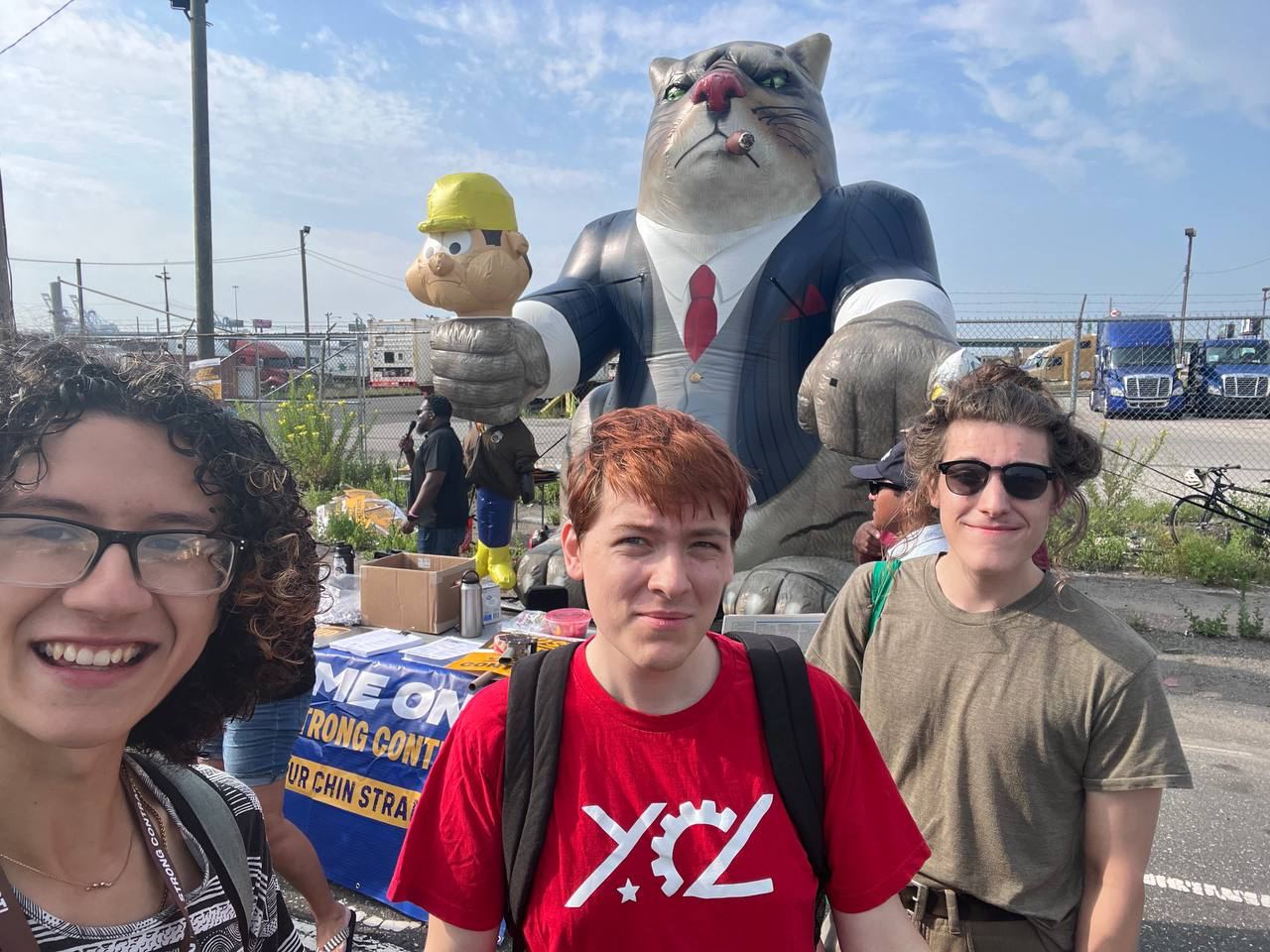 Three people standing in front of a picket line with a 'fat cat' inflatable in the background.