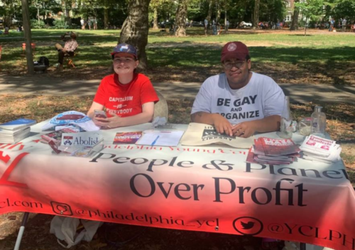 Two people sitting at a folding table in a park. A banner on the table reads 'People & Planet Over Profits'